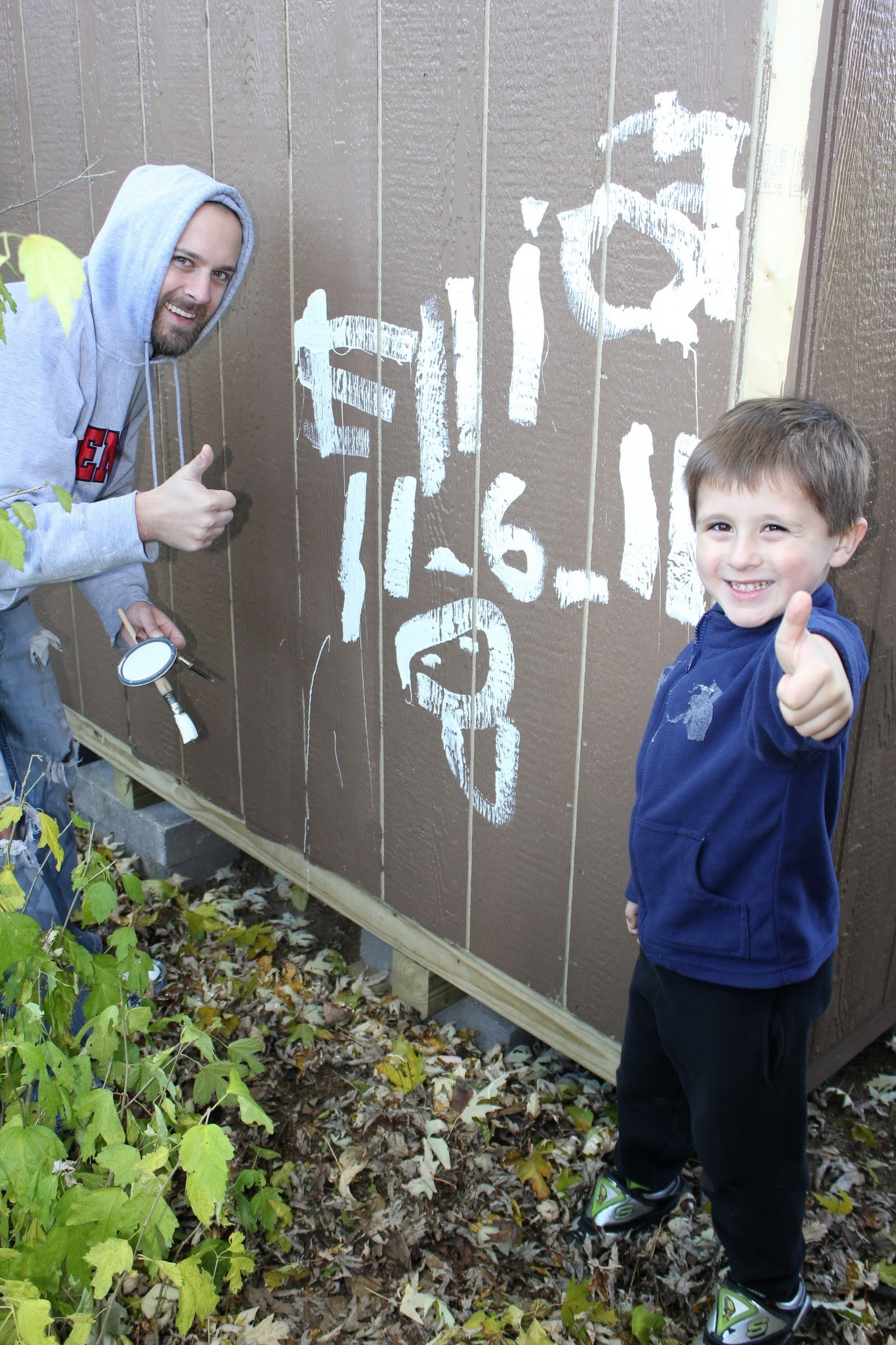 Two people in a playful pose give thumbs up as they stand next to a wall with painted white numbers and letters, surrounded by autumn leaves.