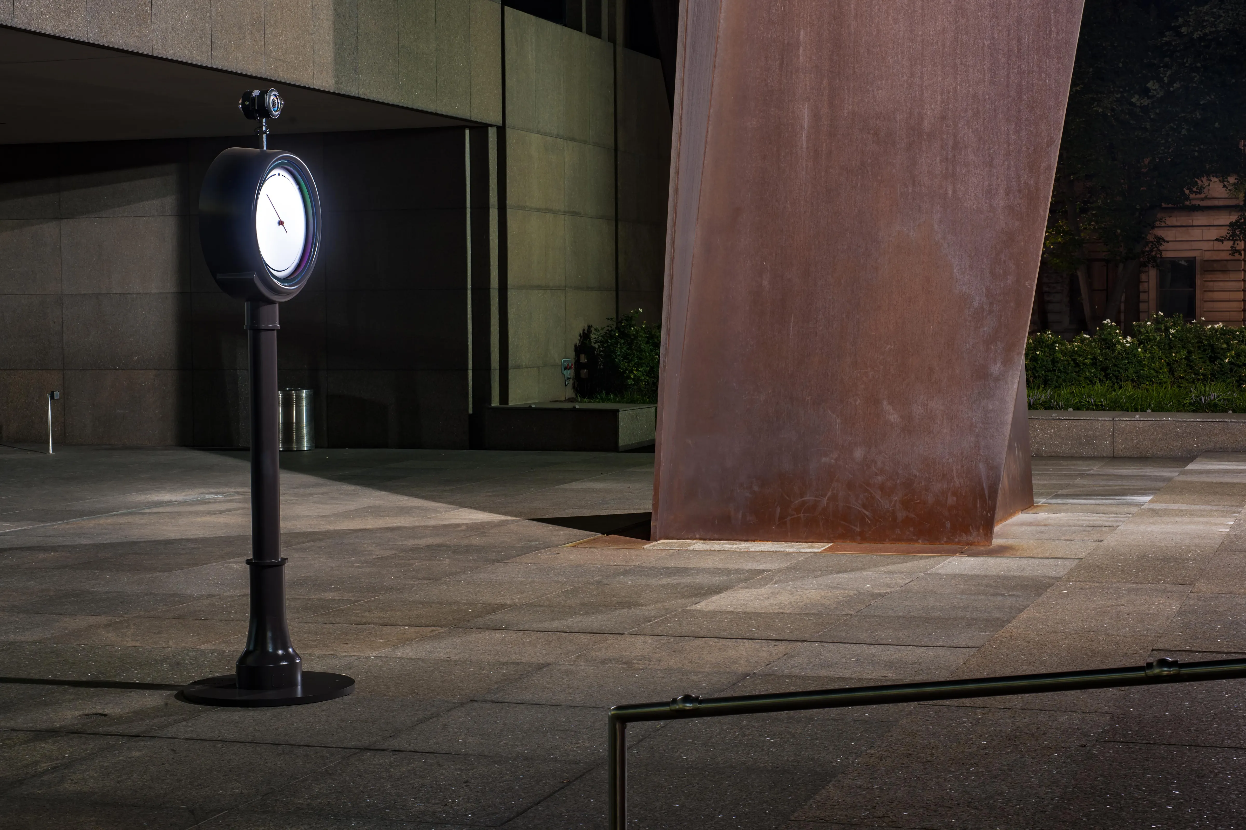 Auto-generated description: A dimly lit plaza features a tall, rust-colored sculpture and a vintage-style clock on a pole.