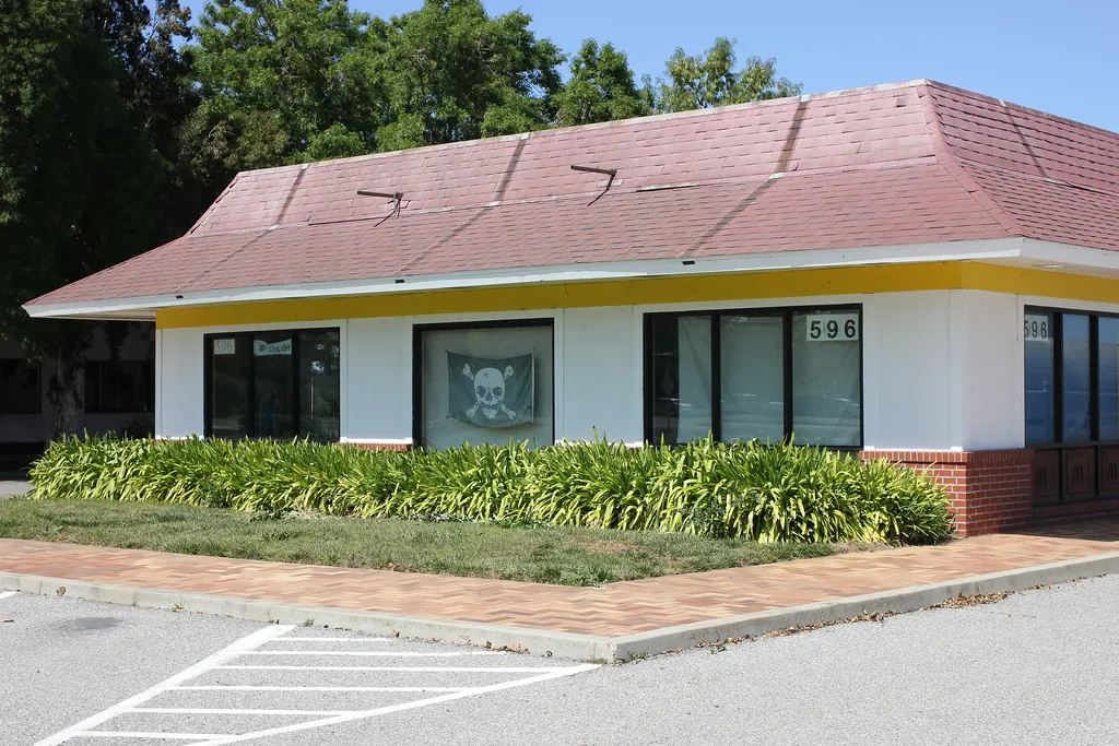 Auto-generated description: A single-story building with a brown roof and a pirate flag displayed in one window is surrounded by greenery and set against a backdrop of trees.