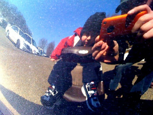 A person is reflected in a shiny surface, crouching down while holding an orange device, with a child wearing a red jacket and sneakers beside them.